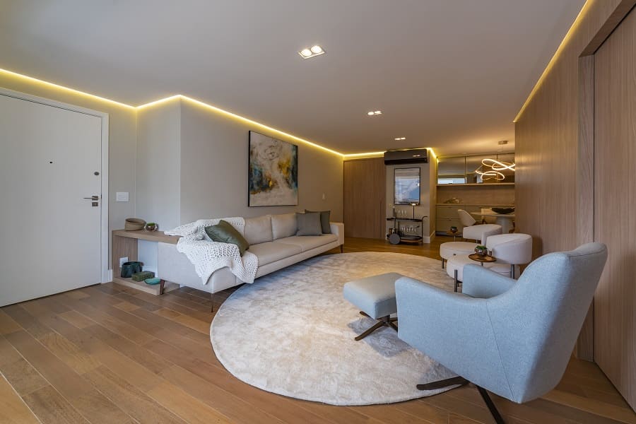 Sala de estar aconchegante com tapete redondo, sofá claro, poltrona azul e painel de madeira natural com iluminação em perfil de LED no apartamento na Vila Olímpia, São Paulo.