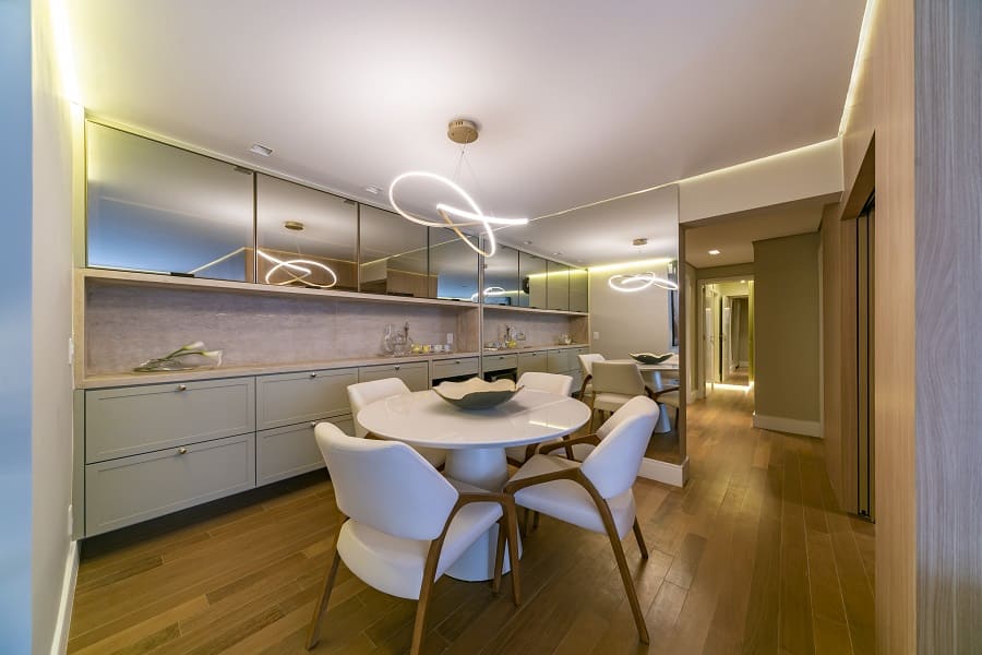 Sala de jantar sofisticada com mesa redonda, buffet com gavetas cinzas, painel espelhado e lustre moderno de LED em projeto de interiores na Vila Olímpia, São Paulo.