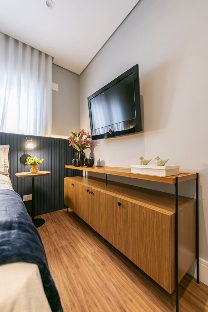 Detalhe da área da TV no quarto de visitas com uma cômoda flutuante de madeira desenhada a 20 centímetros do chão para permitir o armazenamento de malas de viagem. Projeto de interiores no Jardim Caravelas, São Paulo, por Thais Guarizo.