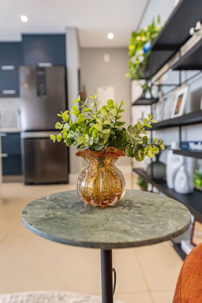 Close em um delicado vaso decorativo de vidro âmbar sobre a mesa lateral de mármore Verde Guatemala, com a cozinha azul integrada desfocada ao fundo. Projeto de apartamento no Jardim Caravelas, São Paulo, por Thais Guarizo.