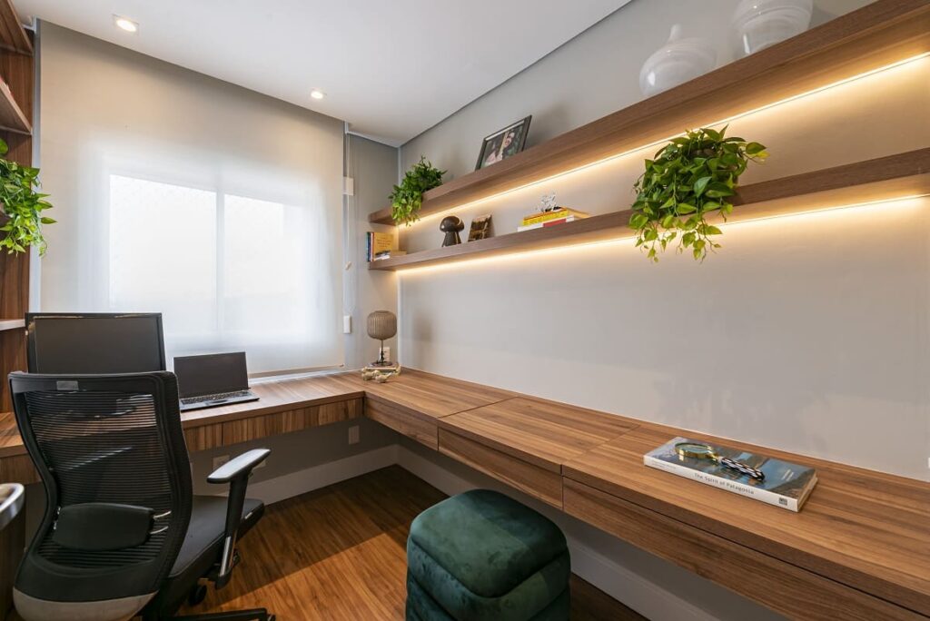 Visão do escritório em casa (home office) destacando a extensa bancada de trabalho em madeira, prateleiras superiores com fita de LED embutida e uma cadeira ergonômica preta.