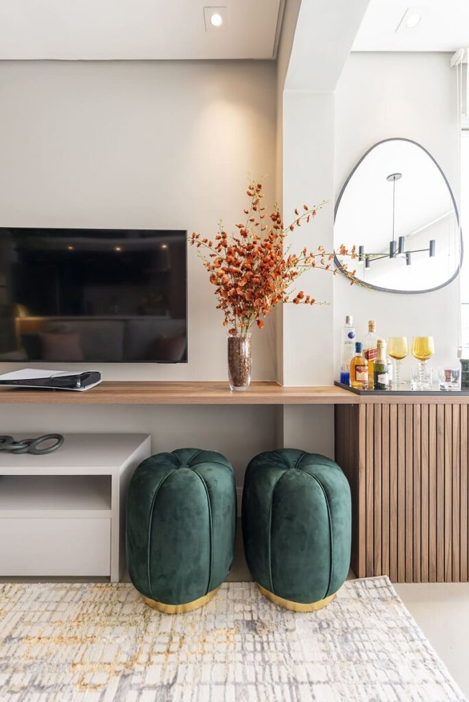 Detalhe da sala de estar focando em dois pufes redondos de veludo verde, um móvel de madeira ripada com adega camuflada, bandeja de bar e espelho assimétrico. Design por Thais Guarizo no Jardim Caravelas, SP.