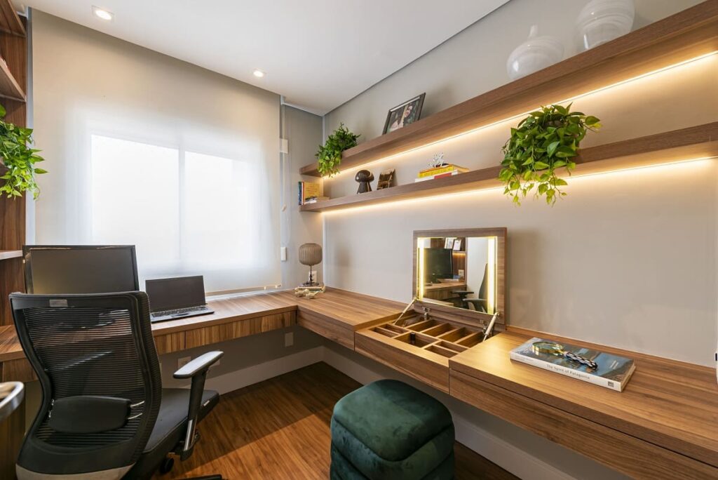 Primeiro plano do home office planejado com bancada em L de madeira e penteadeira camuflada aberta revelando espelho iluminado. Projeto arquitetônico em São Paulo assinado por Thais Guarizo.
