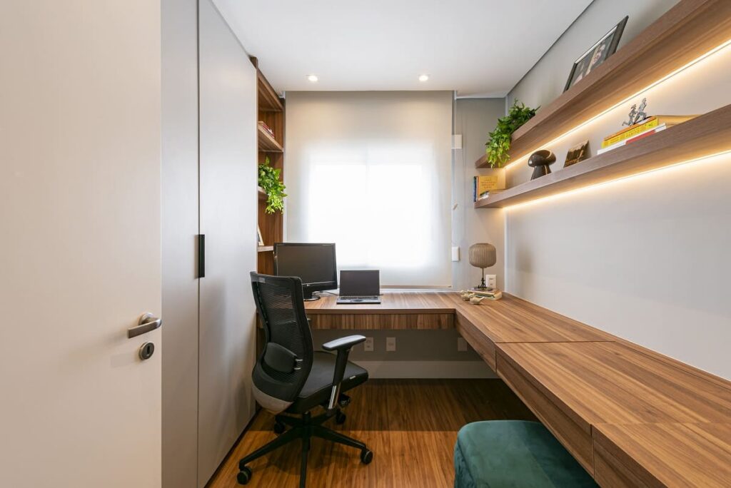 Visão geral do quarto transformado em home office, mostrando a bancada de trabalho sob a janela e um generoso armário planejado cinza para organização. Arquitetura de interiores em São Paulo por Thais Guarizo.