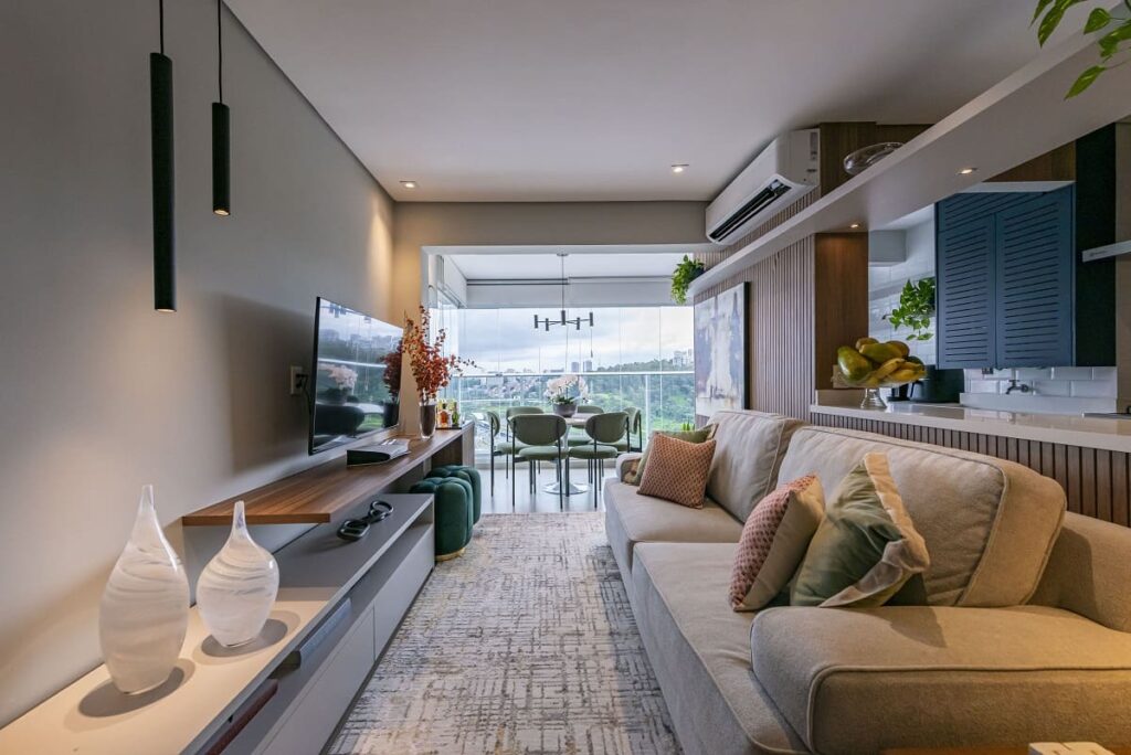 Ampla sala de estar integrada à varanda com vista para o verde, apresentando um sofá cinza claro, tapete texturizado e painel de TV em madeira. Projeto arquitetônico de apartamento no Jardim Caravelas, São Paulo, assinado por Thais Guarizo.