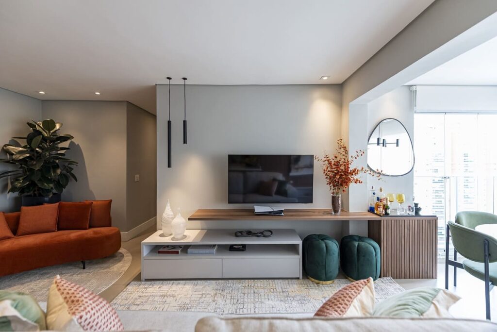Área da TV na sala com rack cinza, prateleira flutuante de madeira com pufes verdes embaixo, e um móvel ripado com área de bar e espelho orgânico. Projeto no Jardim Caravelas, São Paulo, de Thais Guarizo.