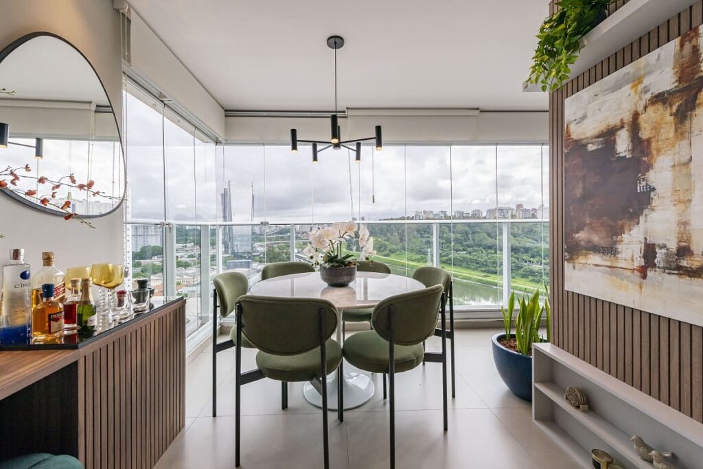 Varanda gourmet com fechamento de vidro, apresentando uma mesa de jantar redonda, cadeiras estofadas verdes e uma bela vista panorâmica para o Parque Burle Marx e a Marginal Pinheiros. Projeto no Jardim Caravelas por Thais Guarizo.