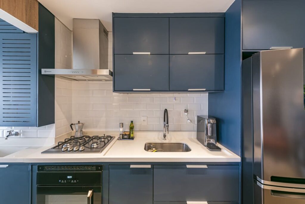 Resultado final da cozinha após a reforma, destacando a elegante marcenaria planejada na cor azul petróleo, bancada clara e revestimento estilo tijolinho branco na parede. Projeto de arquitetura de interiores no Jardim Caravelas, São Paulo, assinado pela arquiteta Thais Guarizo.